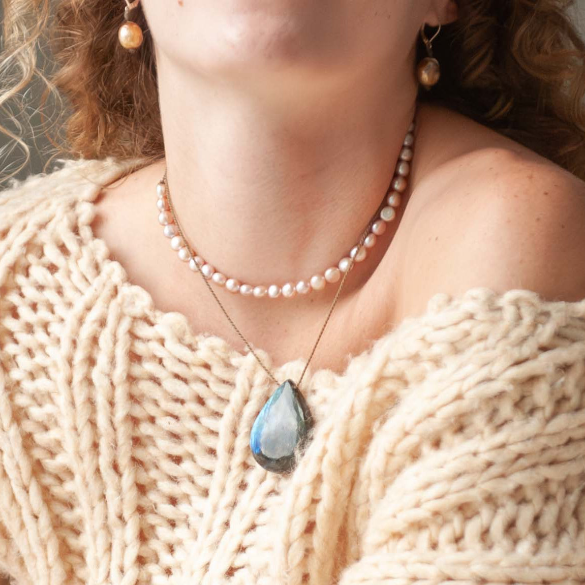 Woman wearing a beige knit sweater and pearl necklace in a cozy room.