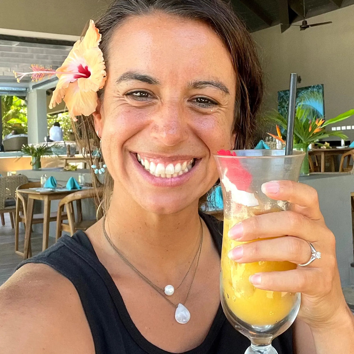 Smiling woman on vacation with a hibiscus in her hair and a tropical drink, wearing the Classic Pearl Necklace layered with a teardrop pendant