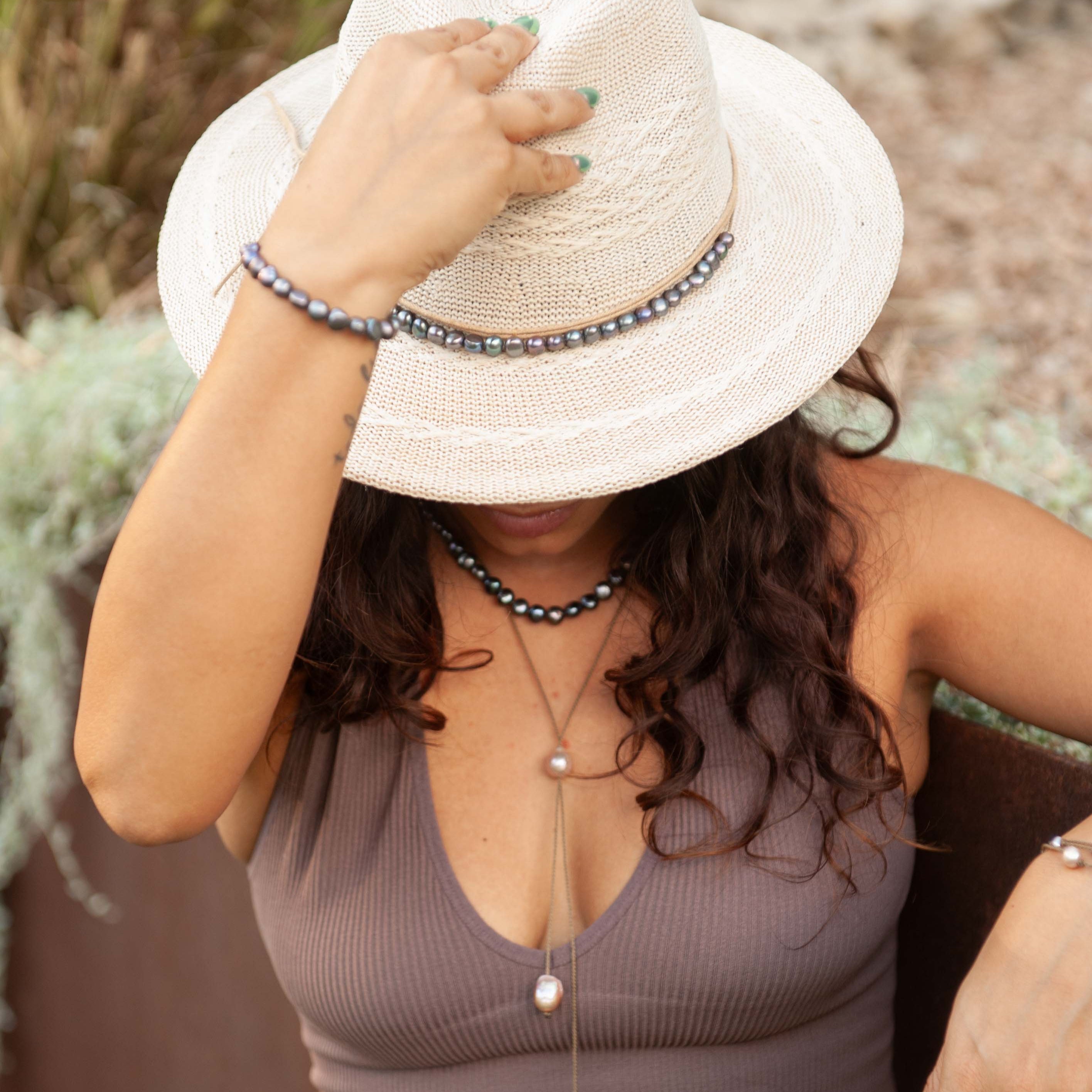 Woman wearing a white hat and a brown top with a pearl necklace, bracelet and hatband standing outdoors.