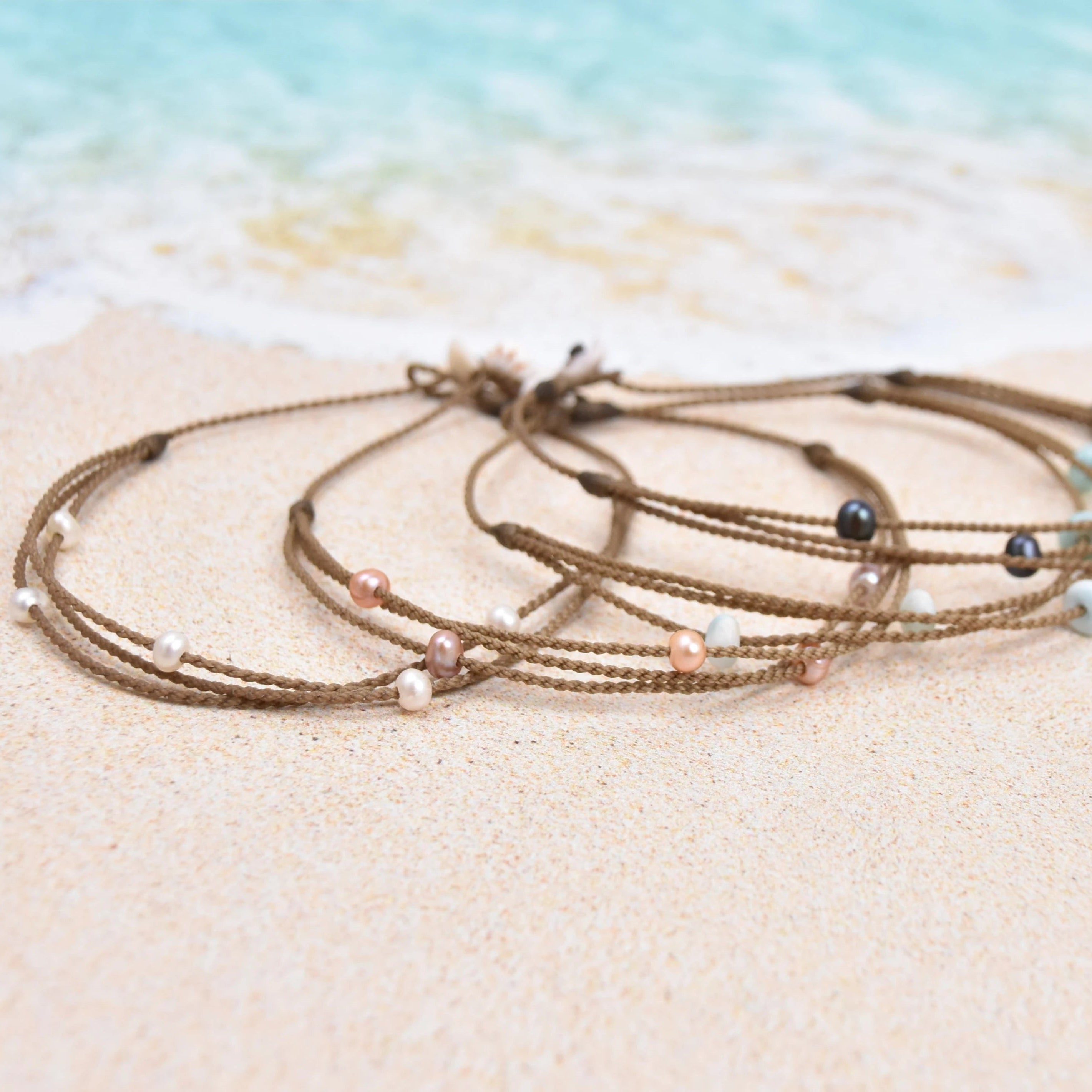 Tula Blue's waterproof Riptide anklet options in white pearls, blush pearls, larimar stones and peacock pearls on the sand by the surf.