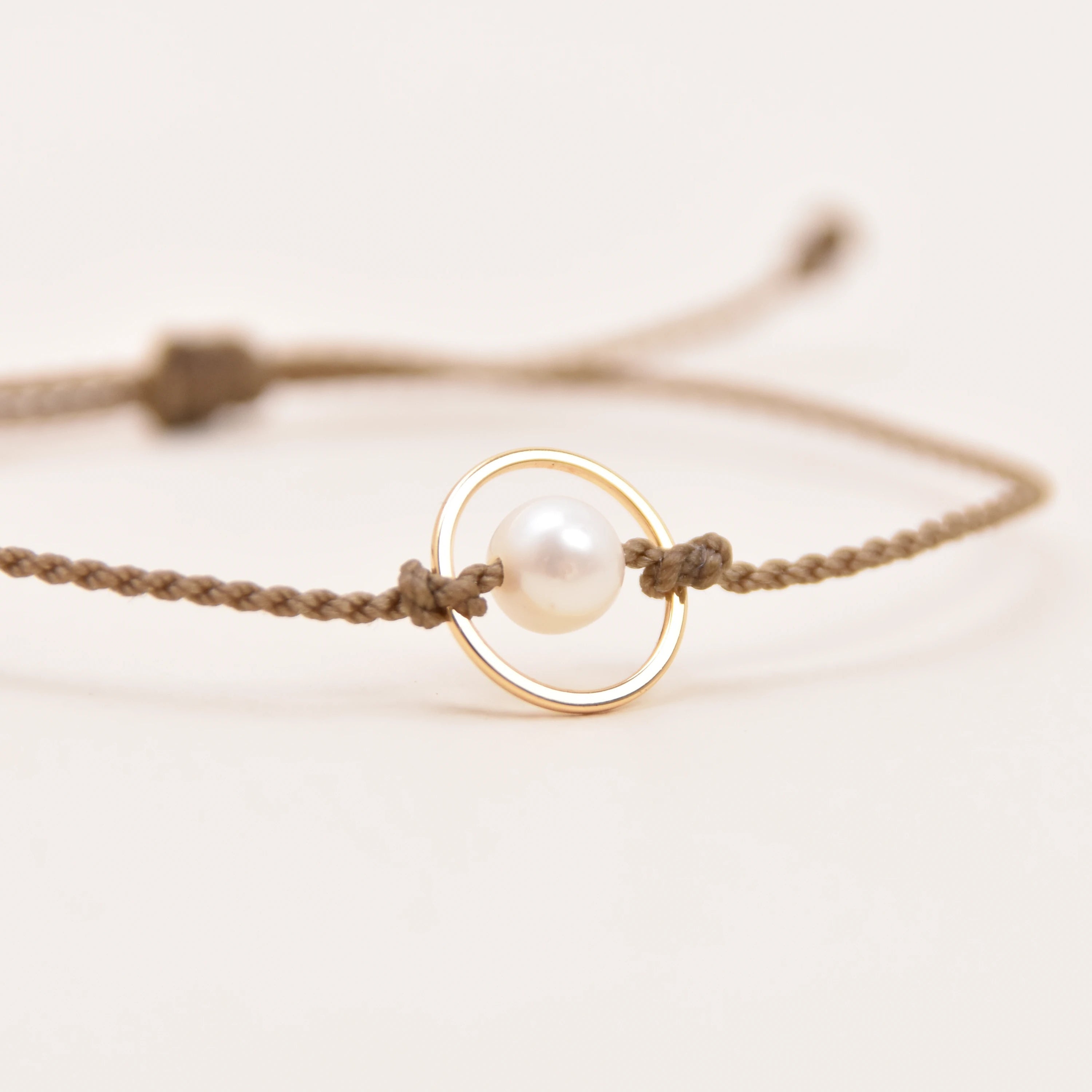 A delicate rope bracelet featuring a central gold ring with a white pearl, photographed on a clean, light off-white background.