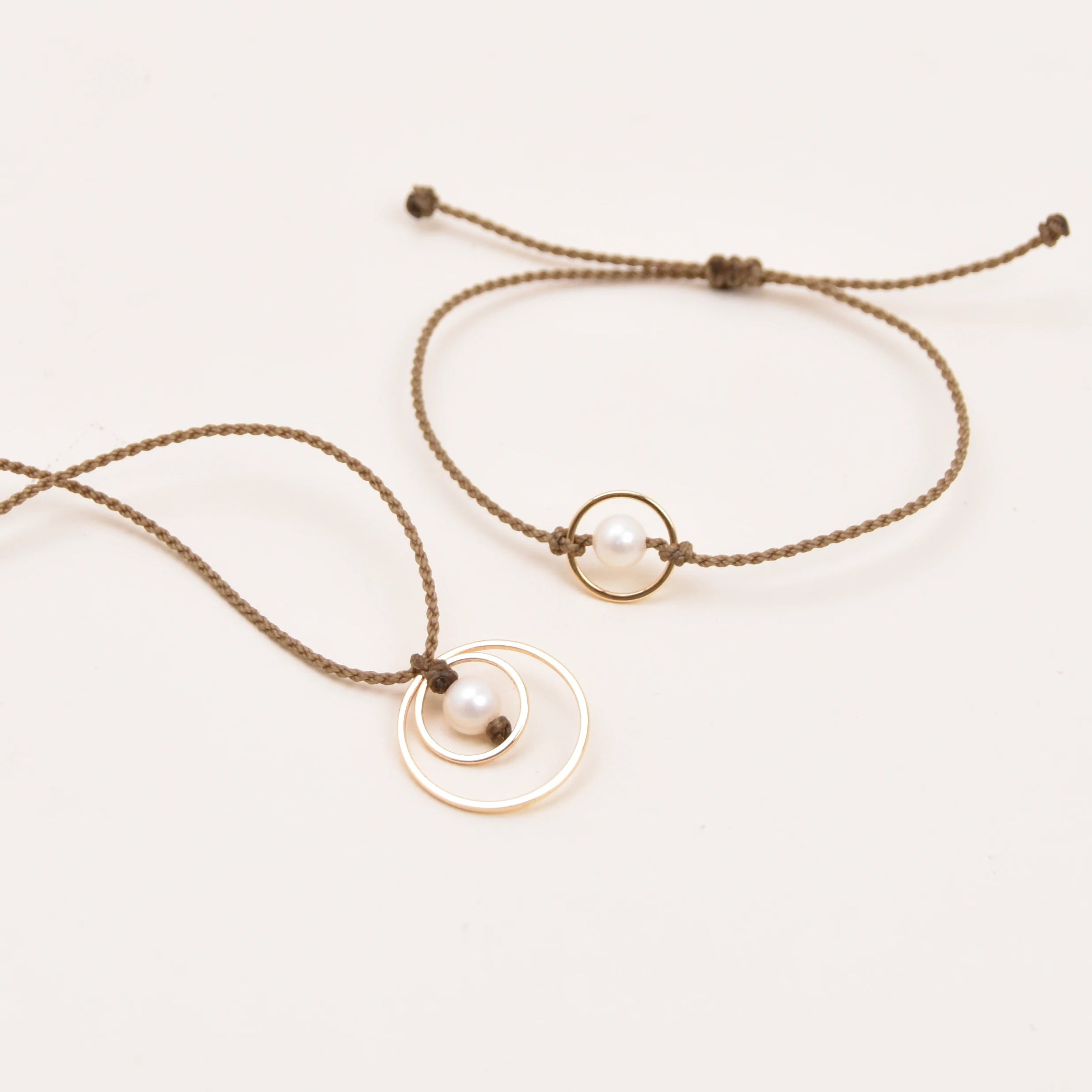 A set of two delicate jewelry pieces on a plain white background: a long necklace of handspun rope, a round gold pendant, and a small white pearl, alongside a matching adjustable rope bracelet with a small circular ring.