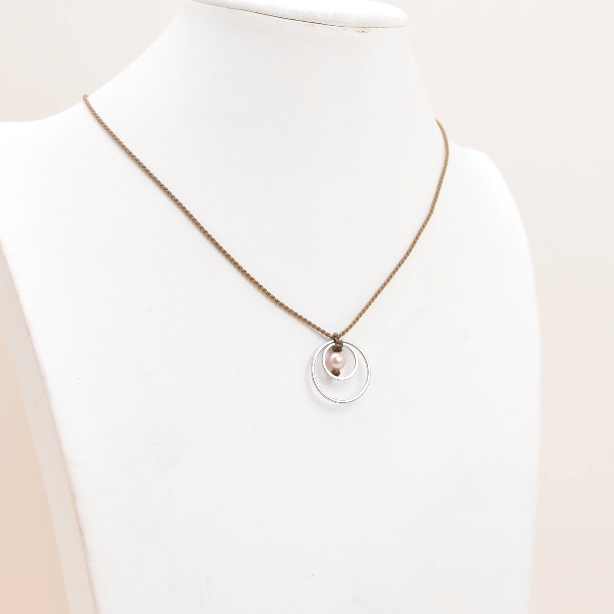 A delicate silver rope necklace with a double-ring pendant displayed on a white jewelry bust against a soft, light beige background.