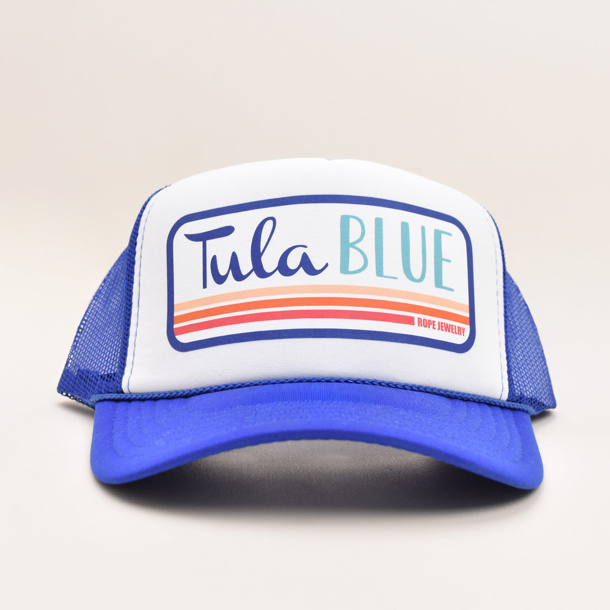 A blue and white trucker hat with a white front panel featuring the text 'Tula BLUE' in blue and teal, with orange and red stripes underneath, set against a plain light background.