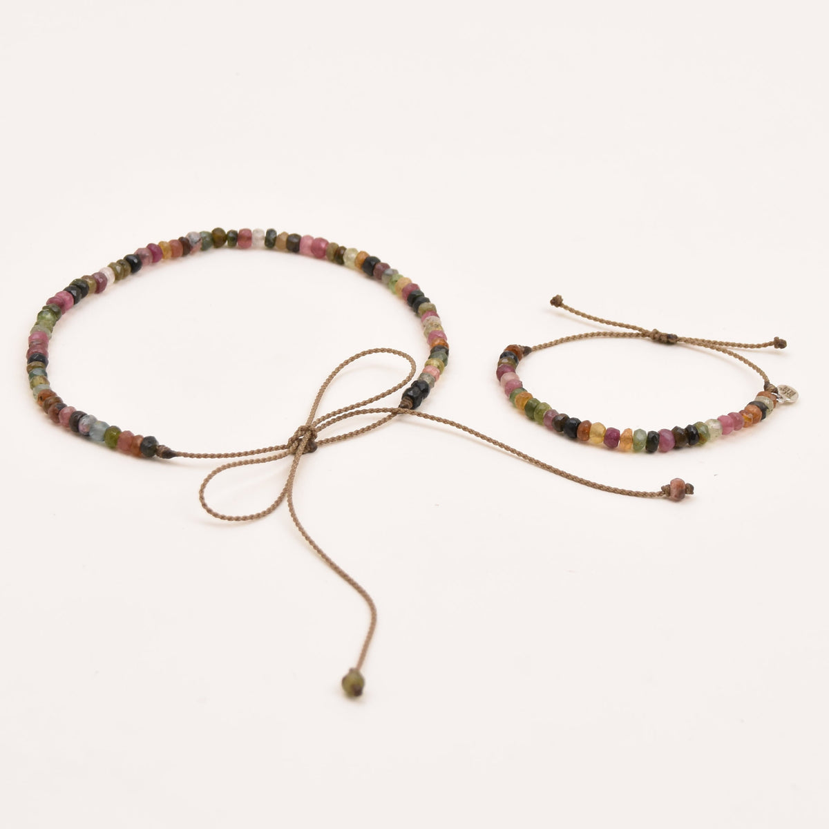 A watermelon tourmaline rope adjustable necklace and matching bracelet laid flat on a plain white background. The jewelry features small multicolored beads with a brown rope and sliding cord closures.