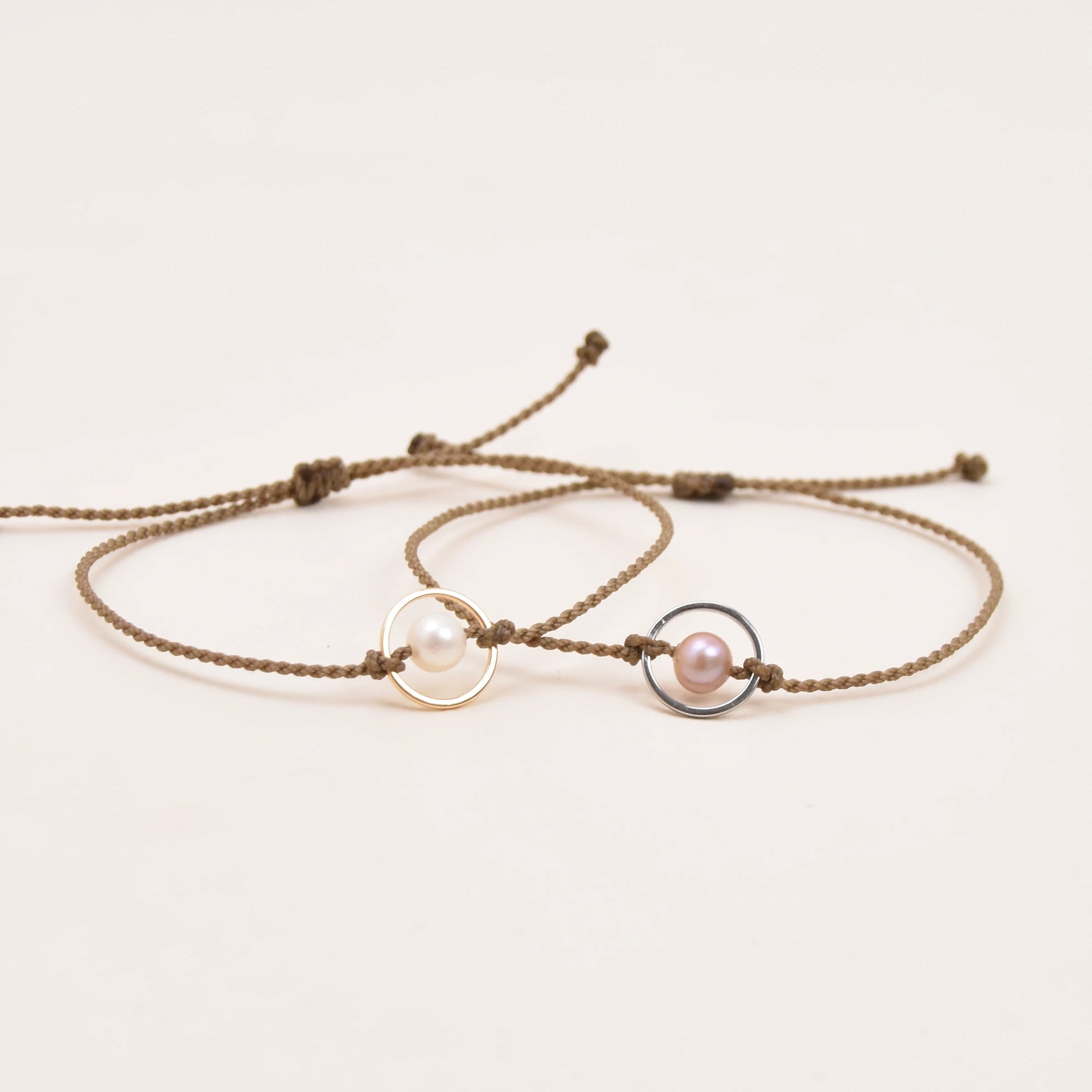 Two delicate adjustable rope bracelets on a plain white background. One bracelet features a polished gold-toned circular disc, and the other features a small pink pearl set in a silver-toned circular frame.