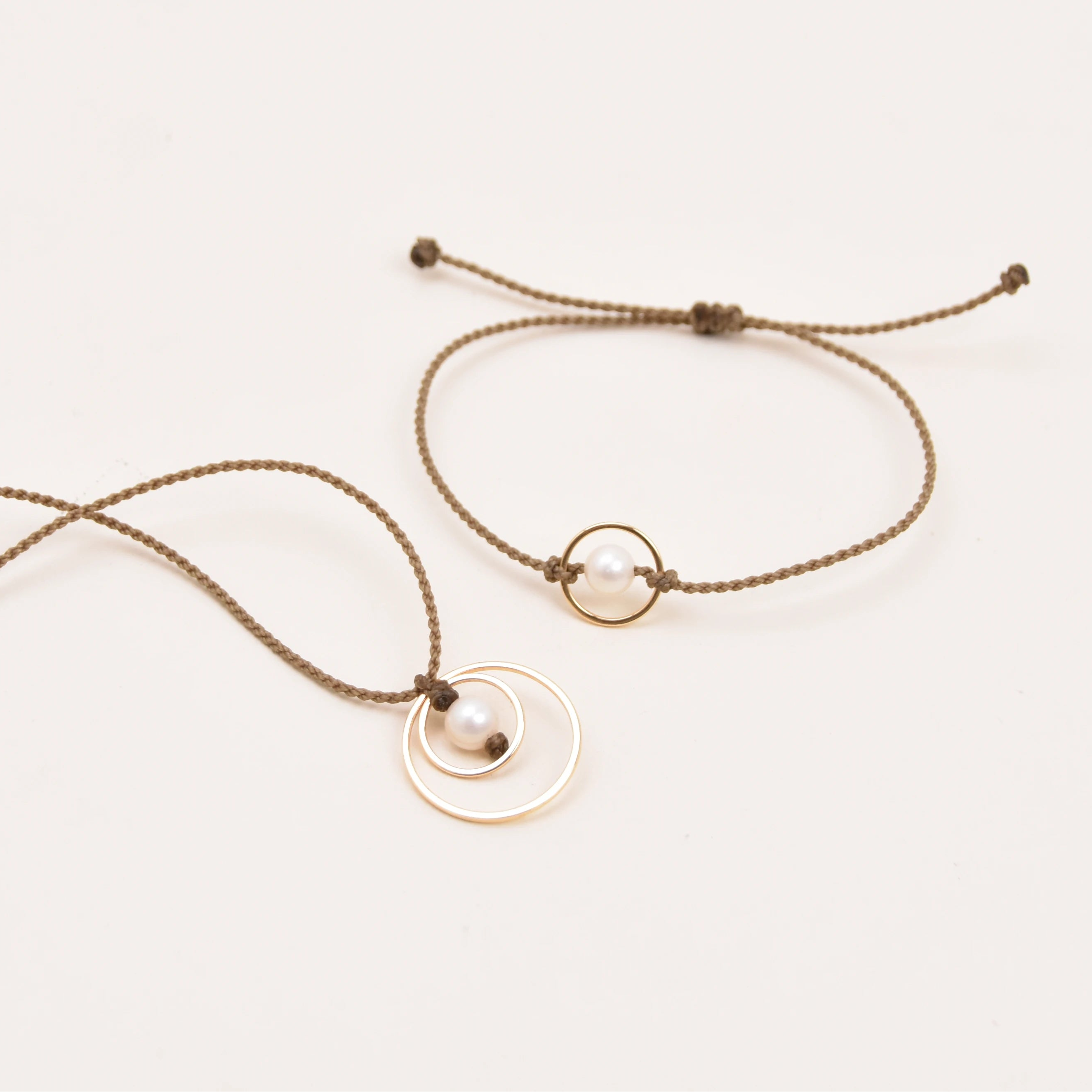 A set of two delicate jewelry pieces on a plain white background: a long rope necklace, a round disc pendant, and a small white pearl, alongside a matching adjustable rope bracelet with a small circular gold ring.