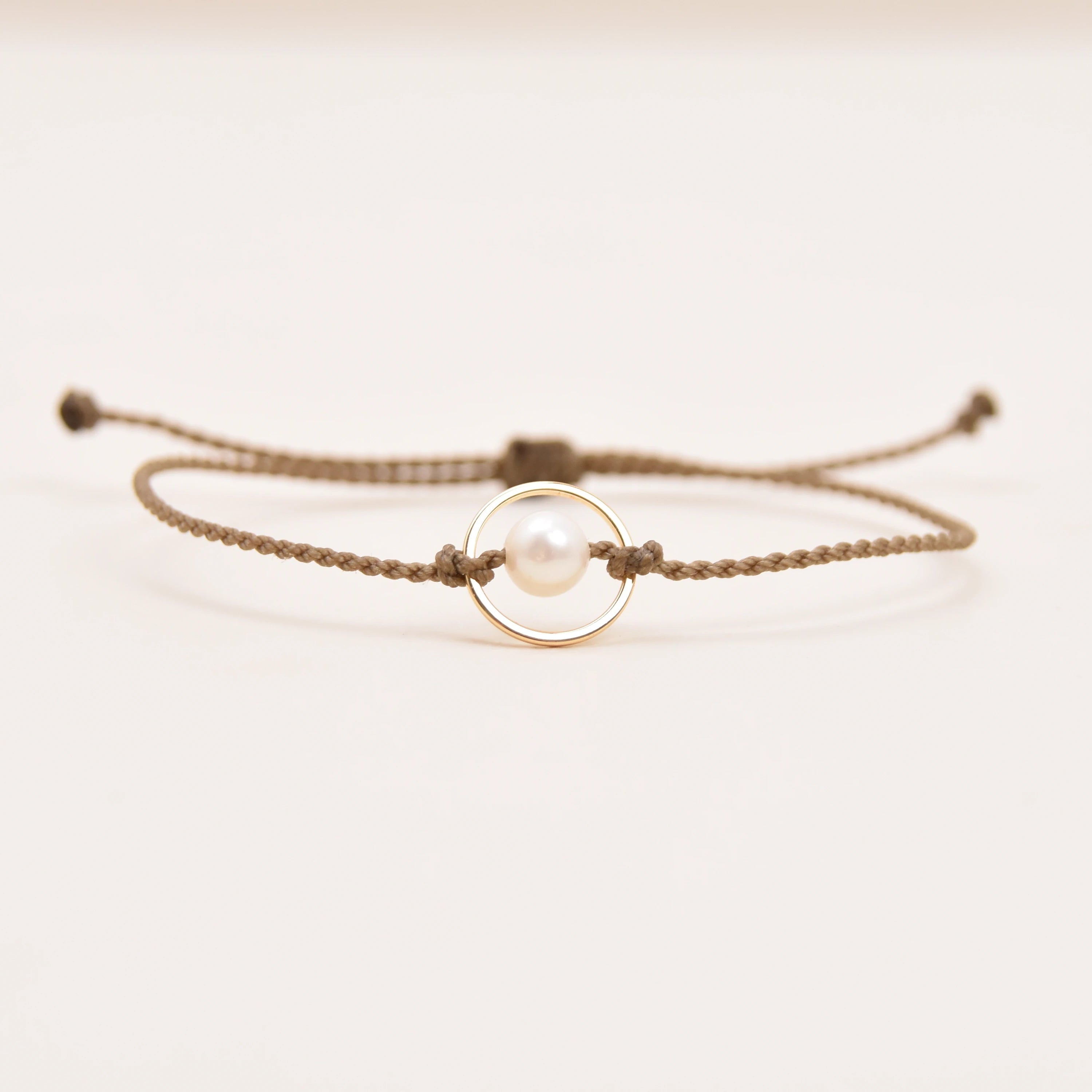 A delicate rope bracelet featuring a central gold ring with a white pearl, displayed on a clean, light off-white background.