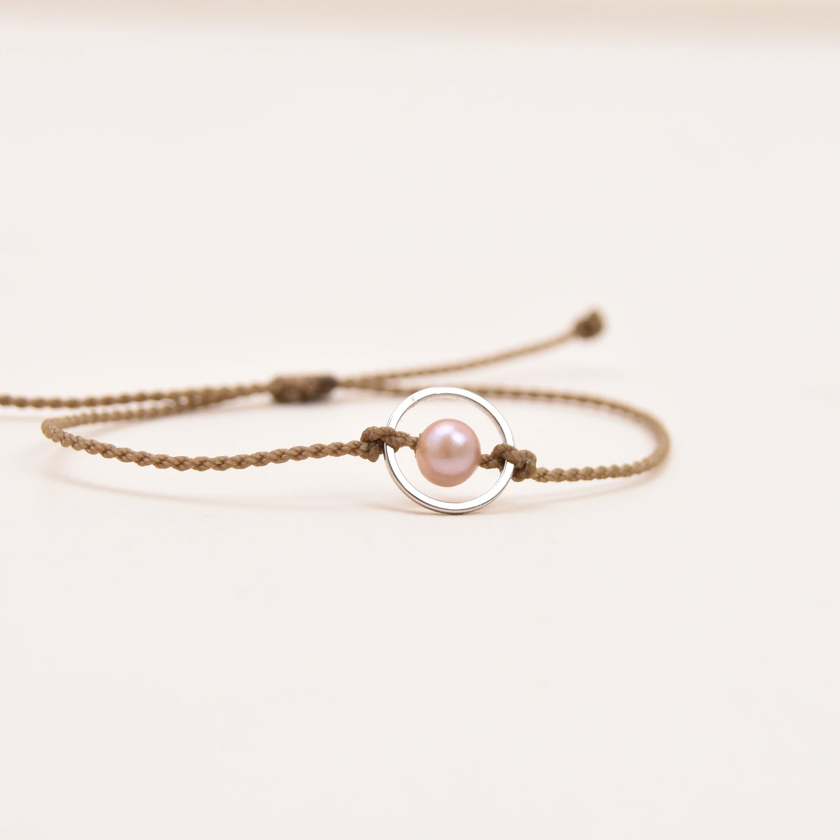 A delicate rope bracelet featuring a central silver circular ring and a small, lustrous pink pearl, displayed on a clean, light off-white background.