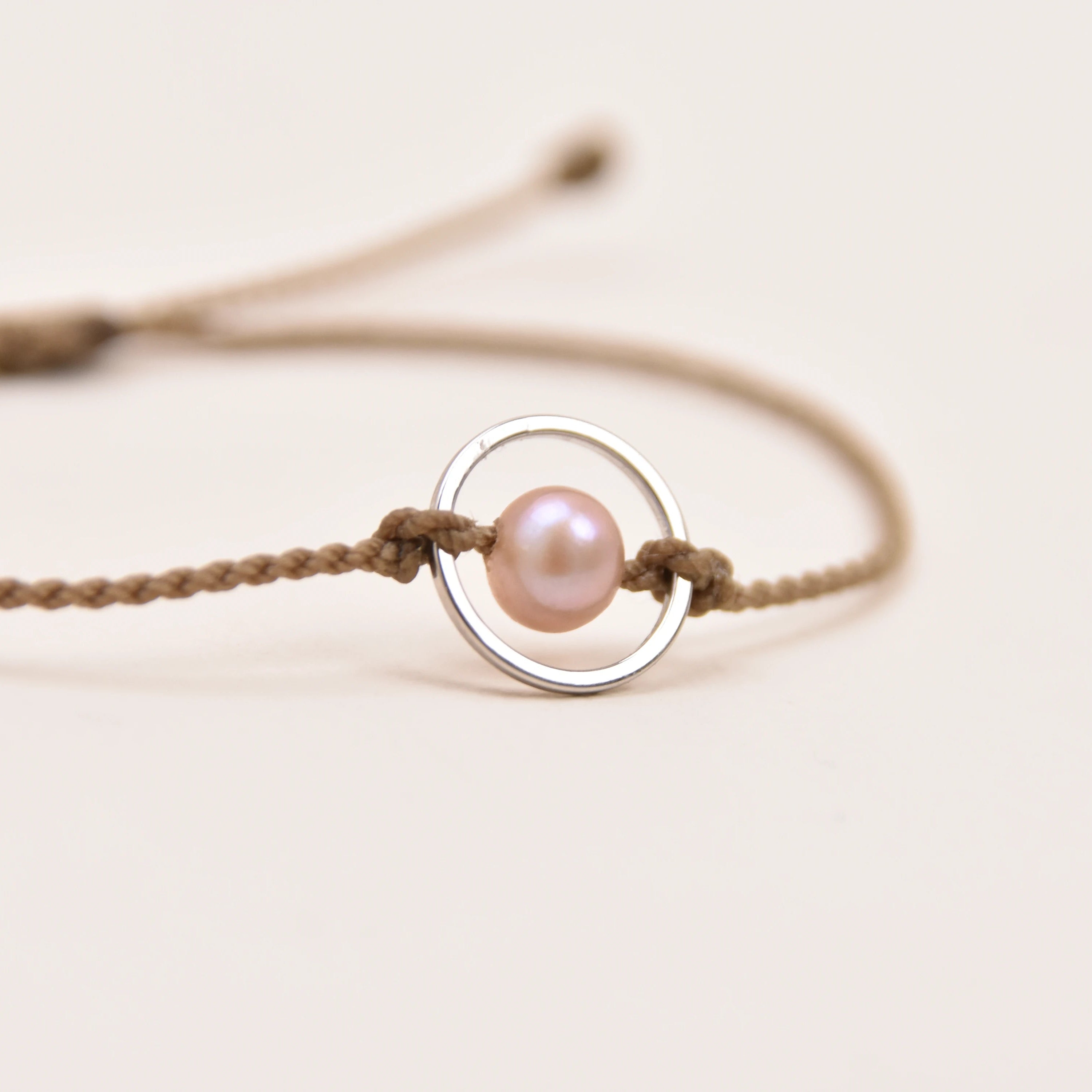 A delicate rope bracelet featuring a central silver ring with a small, lustrous pink pearl bead, set against a plain off-white background.