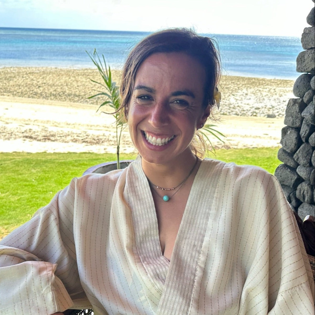 Smiling woman on a beach in Fiji wearing the Classic Pearl Necklace layered between a beaded necklace and a blue stone necklace