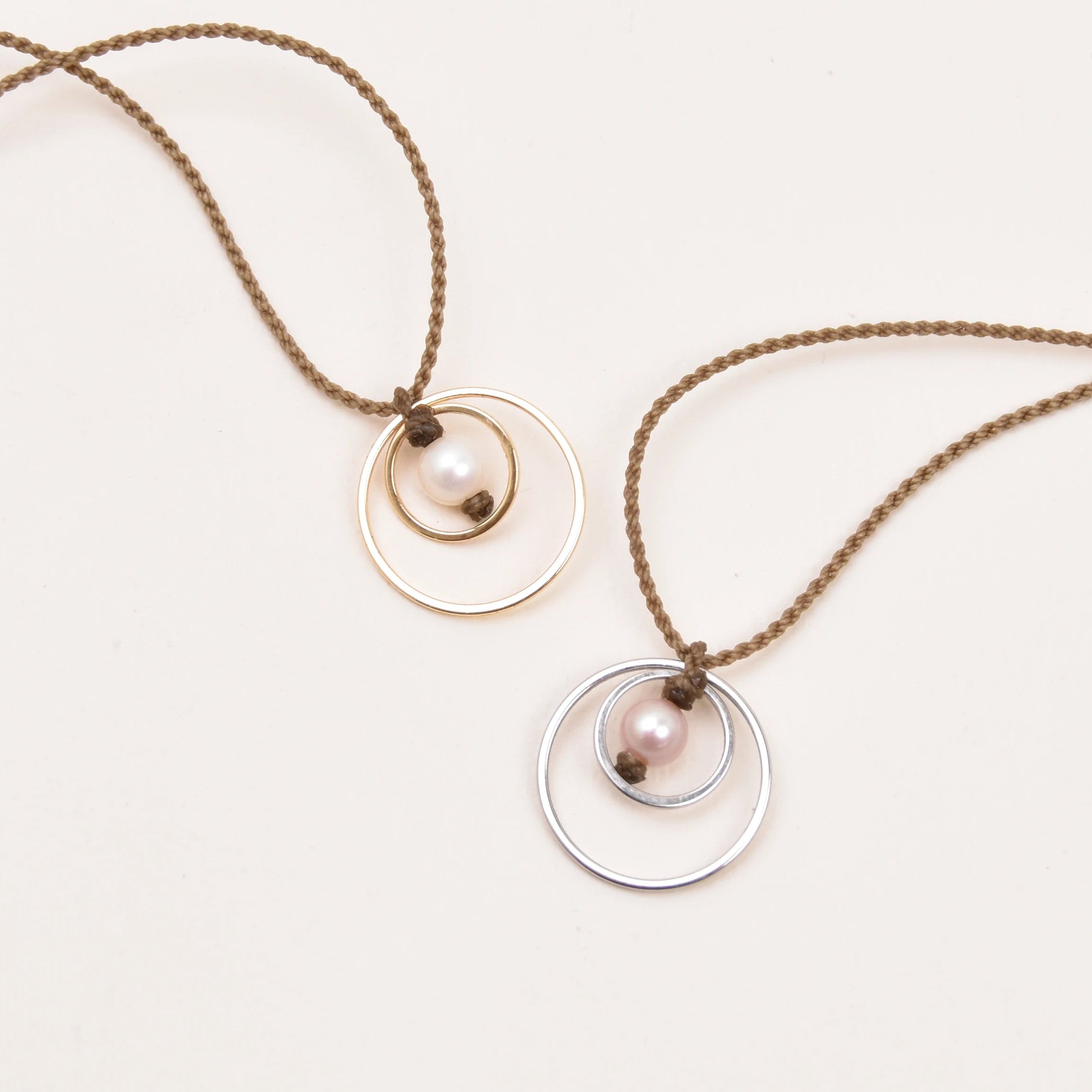Two delicate necklaces with rope, each featuring a small white pearl pendant layered over a thin circular metal frame, one in gold and one in silver, displayed on a clean white background.