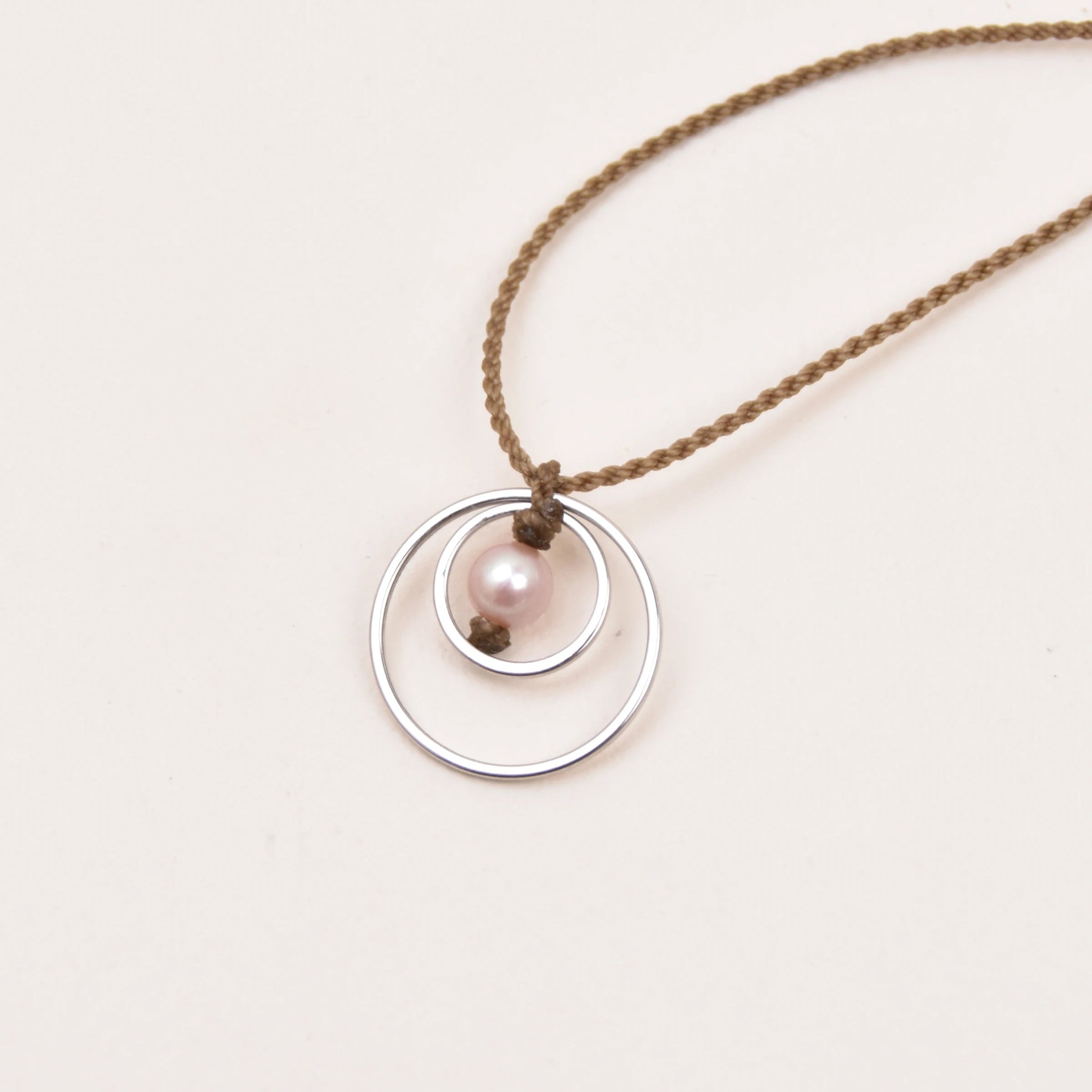 A delicate rope necklace featuring a silver pendant with two interlocking rings and a small pink pearl, displayed on a clean off-white background.