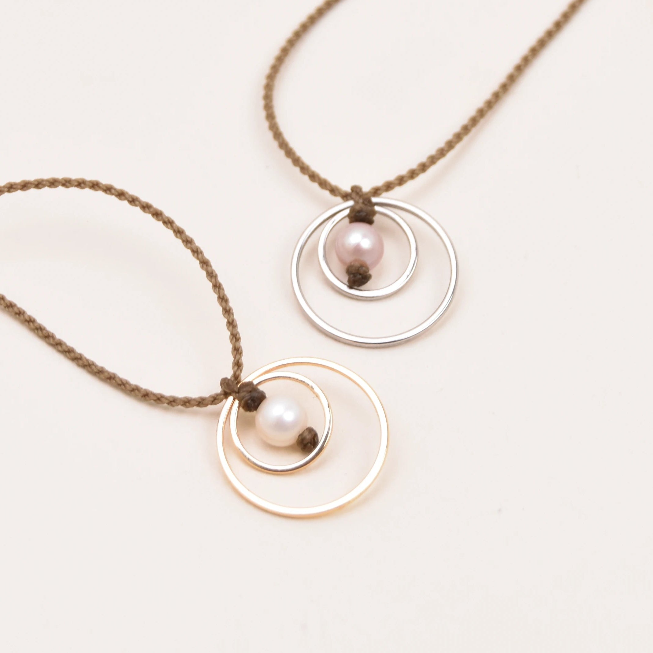 Two delicate rope necklaces and double-ring pendants, one in silver and one in gold, each featuring a small pink pearl, displayed on a clean white background.