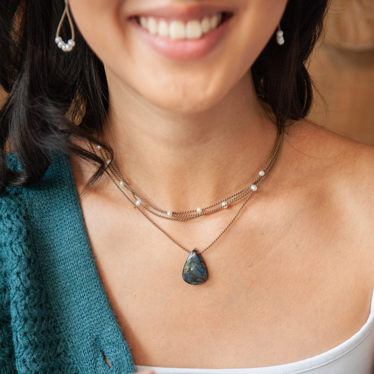 Smiling woman in a teal sweater wearing the Labradorite Gemstone Pendant on hand-spun rope, layered with a white Dainty Riptide Pearl Necklace