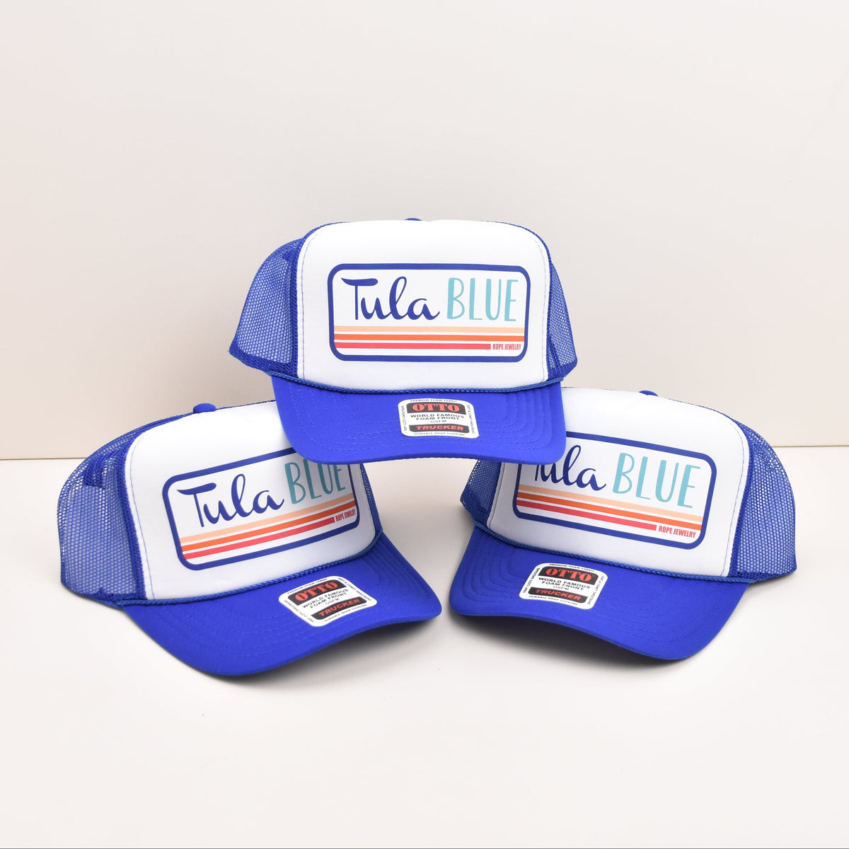 Three blue and white trucker hats with 'Tula BLUE' printed on the front, arranged in a pyramid on a clean white surface against a light beige background.