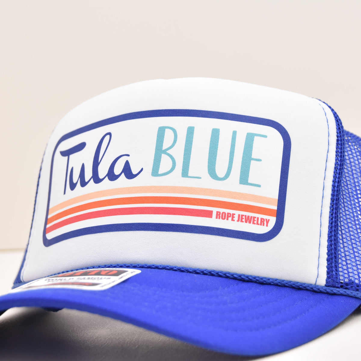 A blue and white trucker hat with a white front panel and blue mesh back. The front features a graphic patch that reads 'Tula BLUE' with orange and red stripes and 'ROPE JEWELRY' in red text.