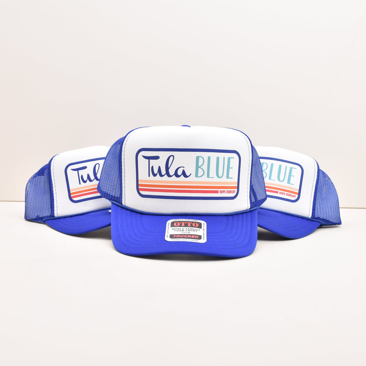 Three blue and white trucker hats with 'Tula BLUE' printed on the front, arranged on a light beige studio background.