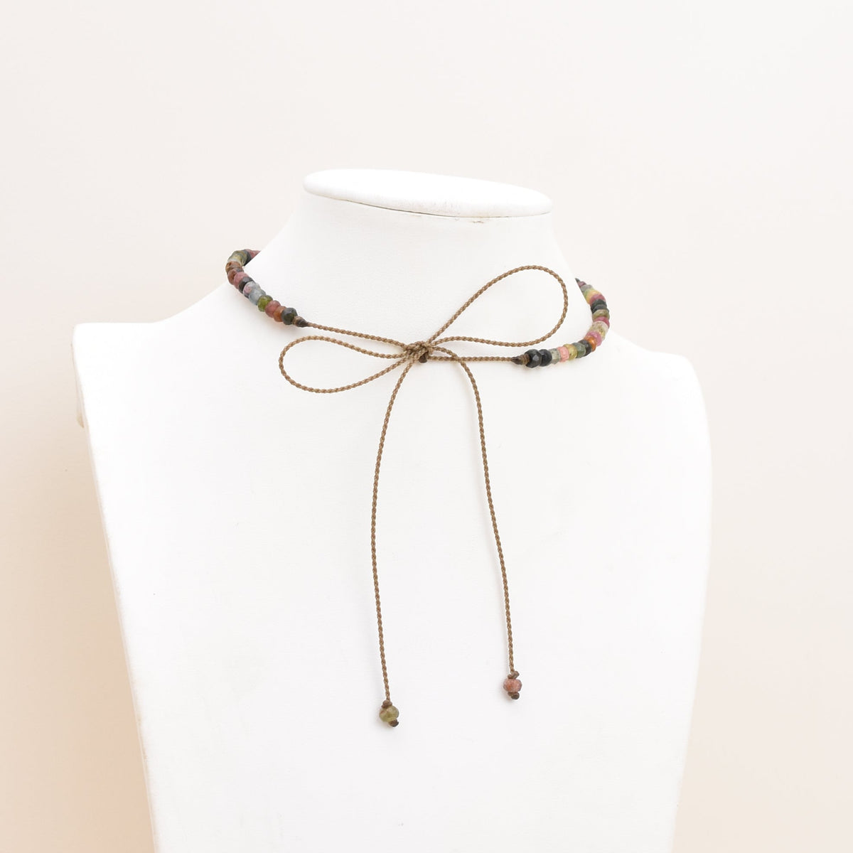 A delicate rope choker necklace with small dark tourmaline rondelles and dangling tips, displayed on a white mannequin bust against a soft beige background.