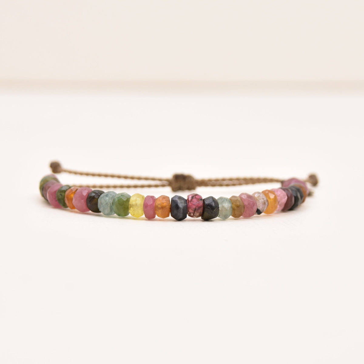 A rope cord bracelet with small, faceted gemstones (watermelon tourmaline), including pink, yellow, green, blue, and purple, on a plain white background.