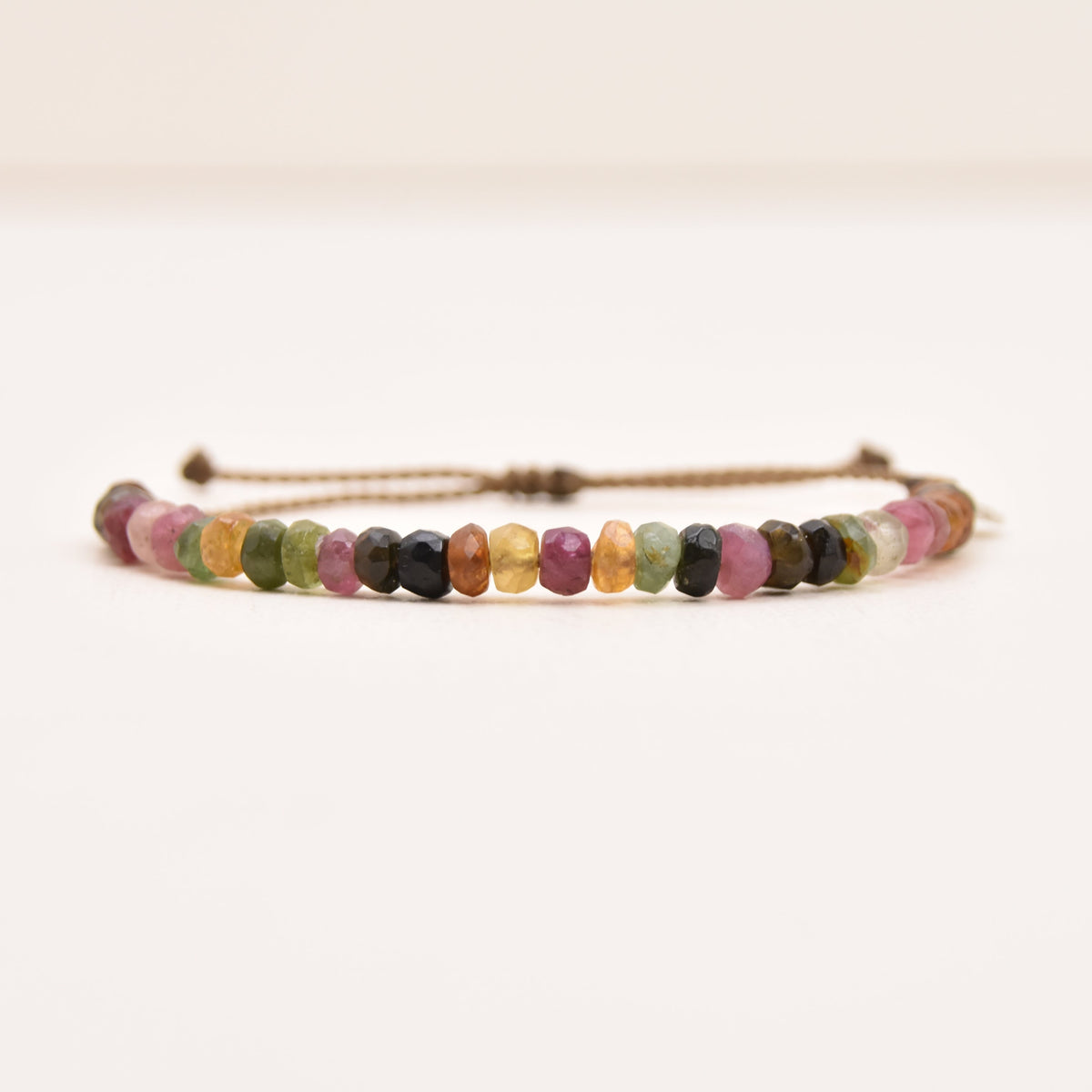 A watermelon tourmaline adjustable rope bracelet featuring a series of small, multicolored tourmaline rondelles on a light brown cord, displayed against a plain off-white background.